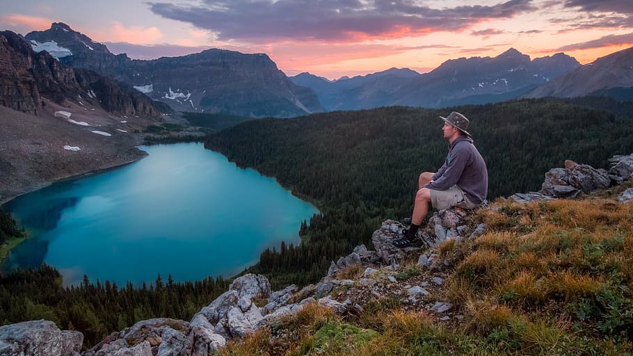 man sitting on rocky hillside watching lake near mountain during daytime, HD wallpaper