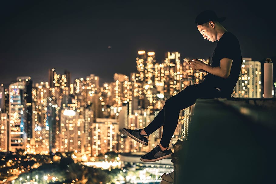 photo of man in black cap, T-shirt, pants, and low top sneakers sitting on edge with high rise buildings with lights during night time HD wallpaper