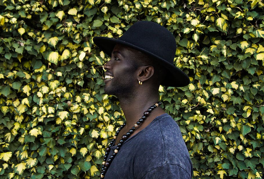 man standing beside living wall, smiling man wearing beaded necklace and black hat standing near yellow and green leaf plant HD wallpaper