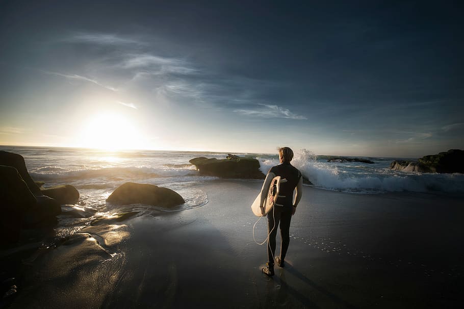 man standing carrying surfboard near seashore during daytime, man holding bodyboard standing facing seashore HD wallpaper