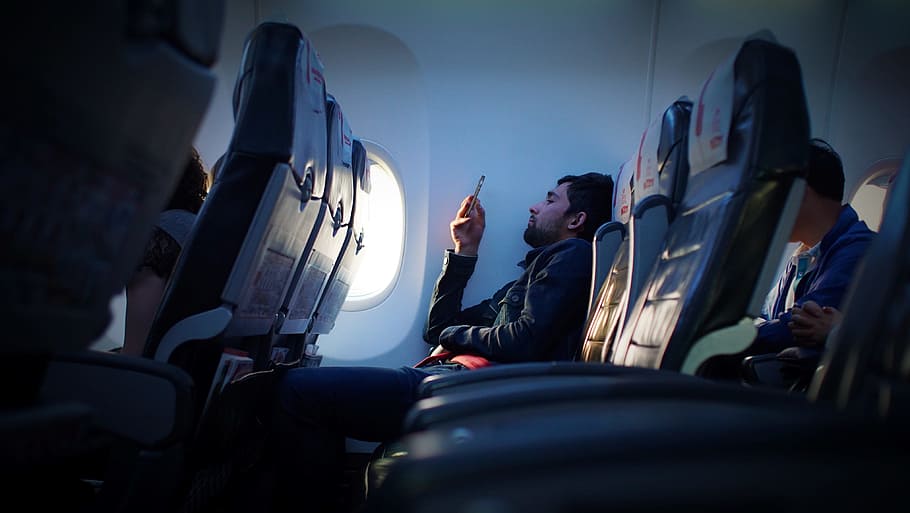 person sitting inside airplane using smartphone, man wearing black dress shirt holding phone in the airplane seat