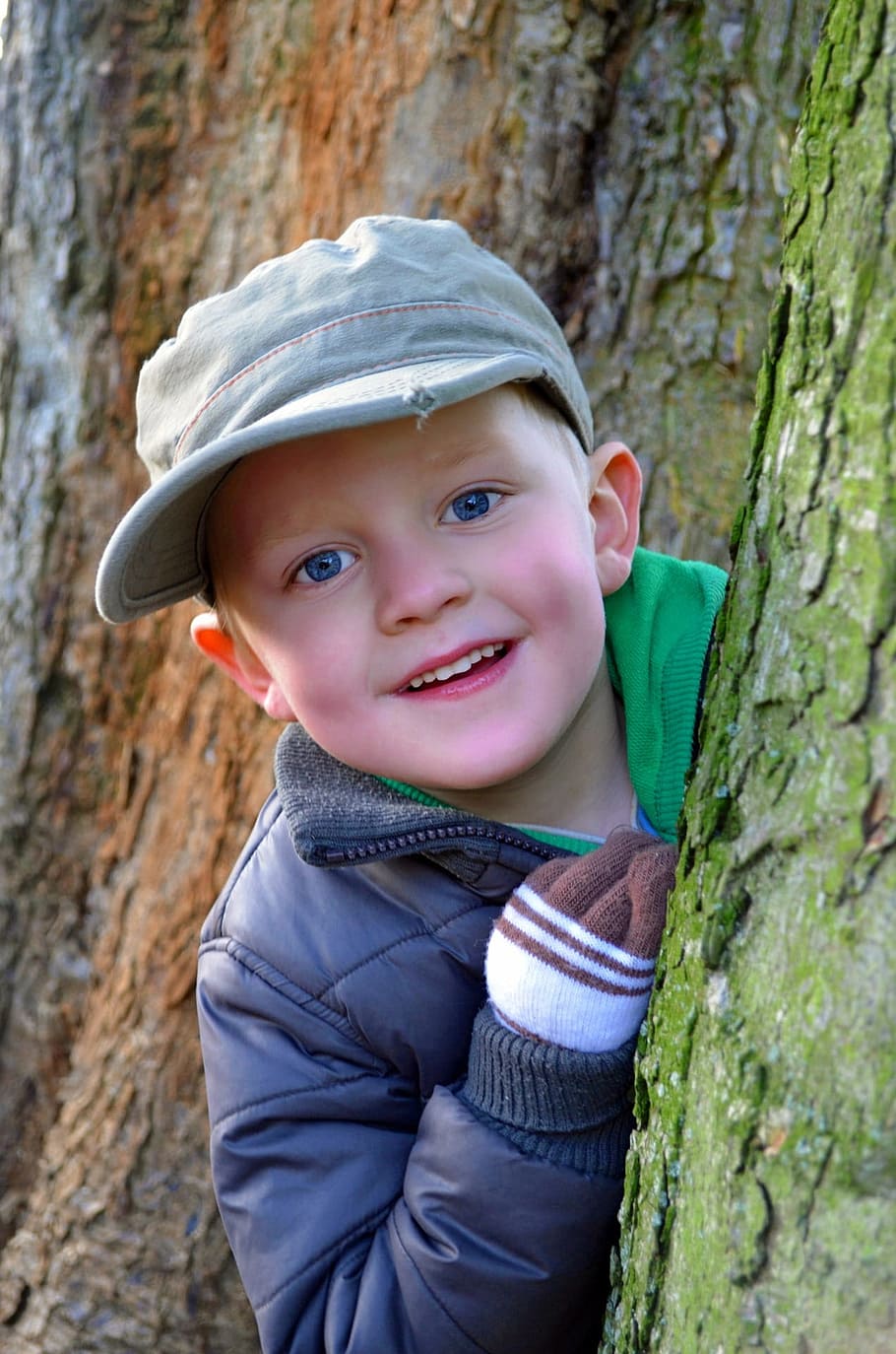 smiling boy wearing gray cap and jacket peeking beside tree trunk with moss