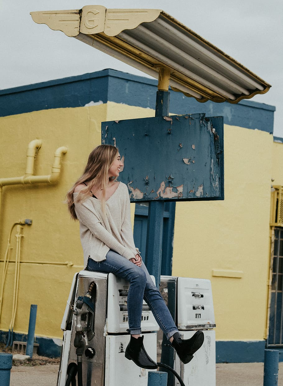 woman sitting on white gasoline pump machine during daytime, woman sitting on fuel dispenser