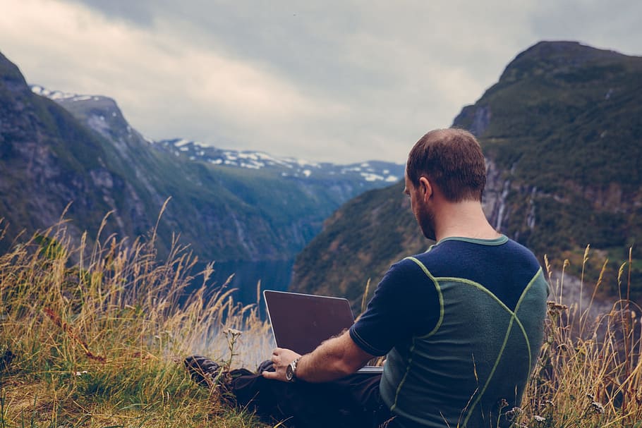 man holding laptop sitting on grass field facing valley with river HD wallpaper
