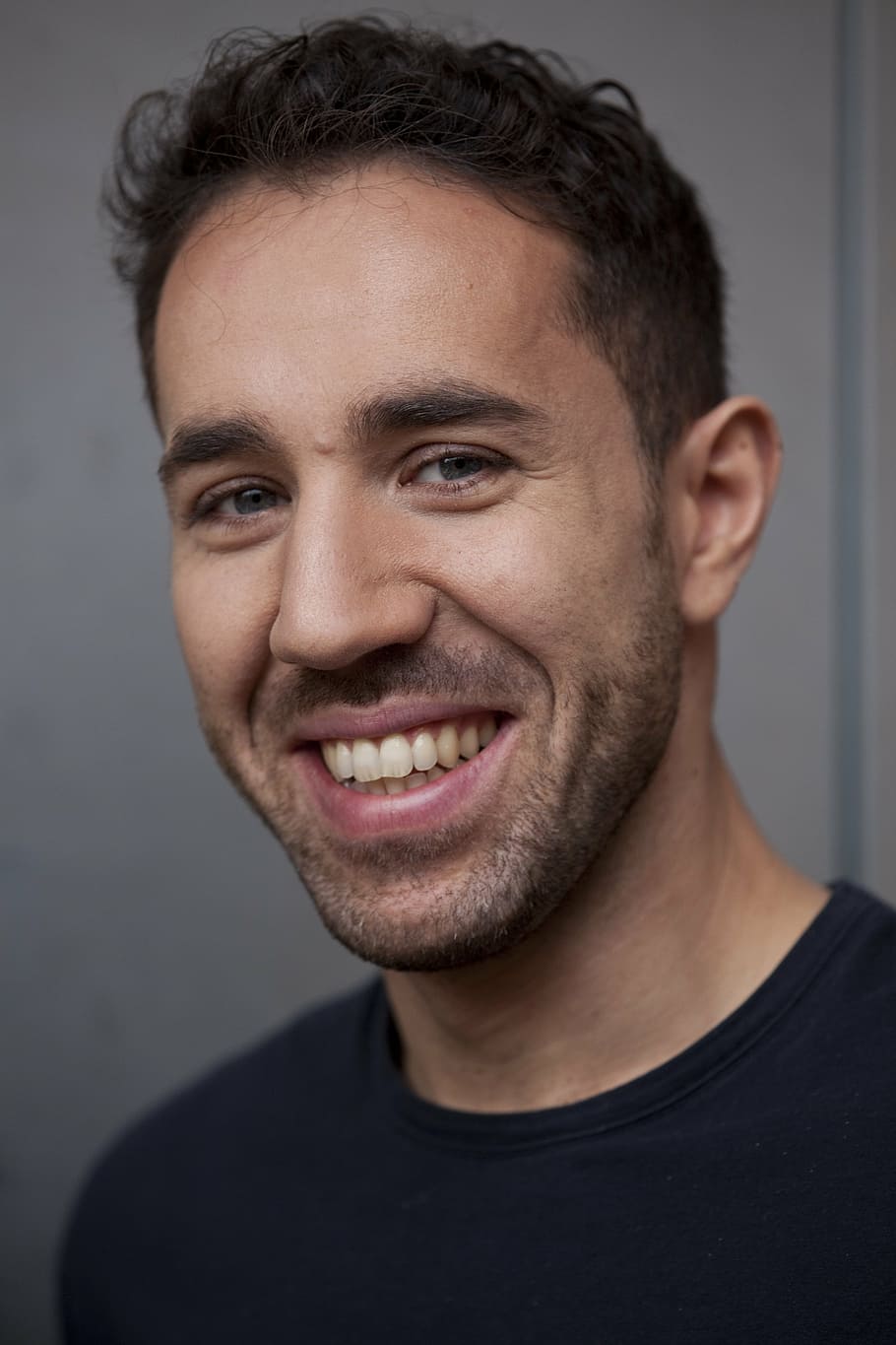 smiling man in black crew neck shirt beside a white wall, face HD wallpaper