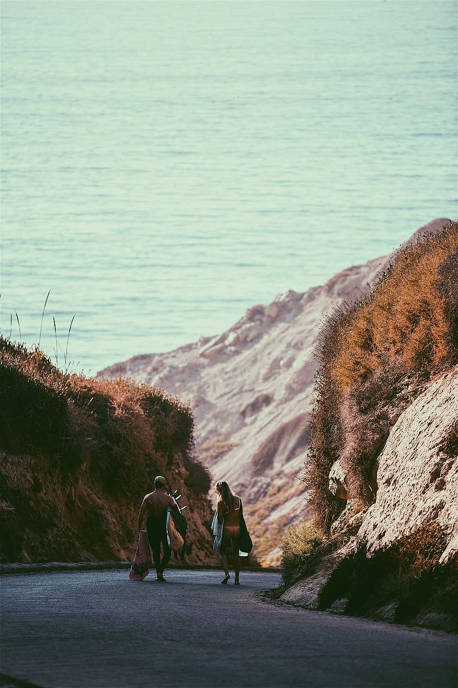 man and woman walking near rock formation, man and woman walking on road towards body of water HD wallpaper