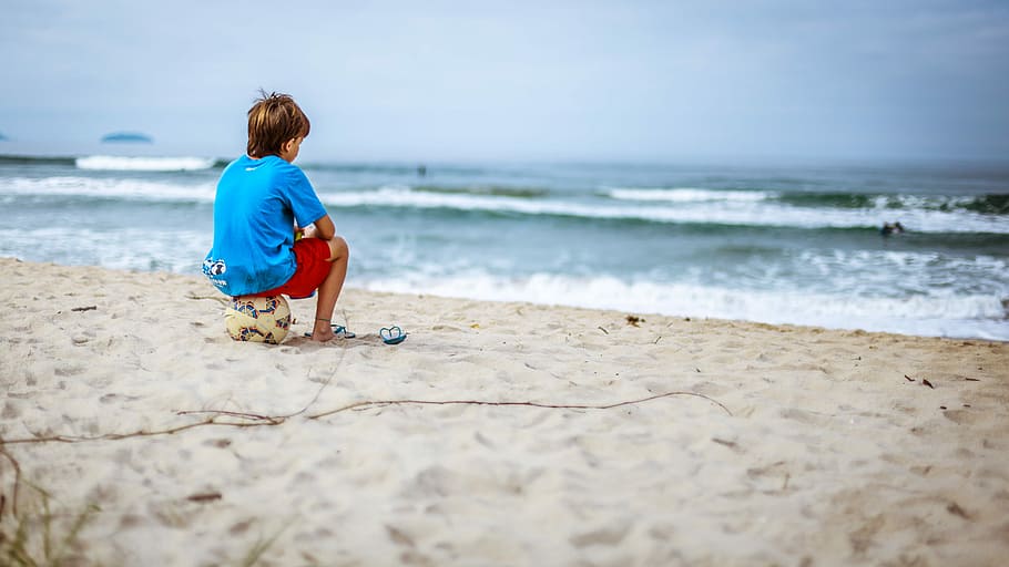 boy sitting on ball while staring at the ocean, child in shore sitting on soccer ball HD wallpaper