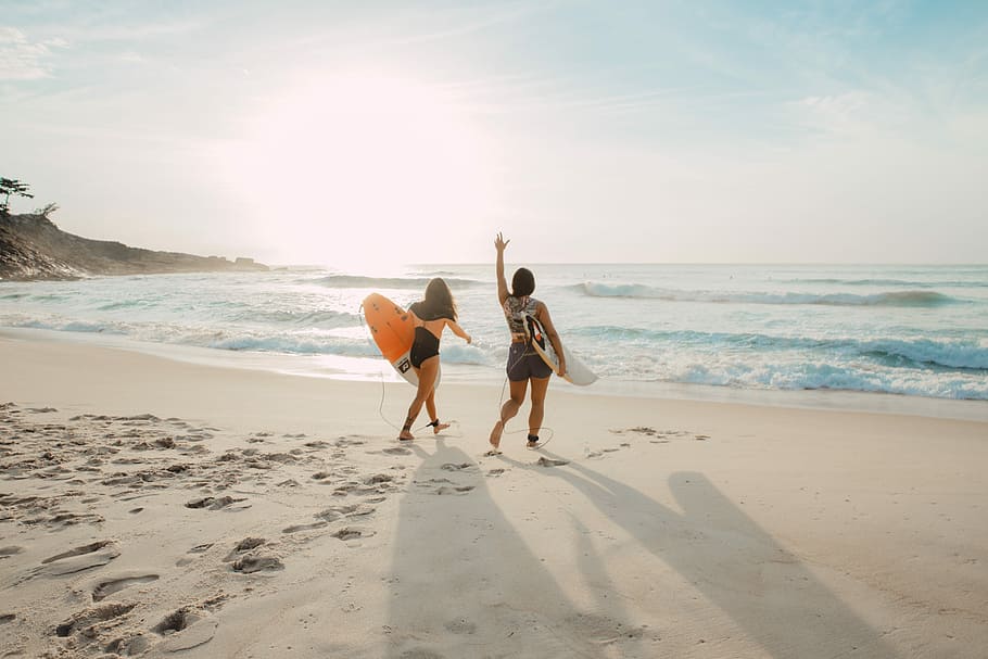 two women walking towards the ocean carrying surfboards during day, two woman walking on seashore with surfboard on hands HD wallpaper