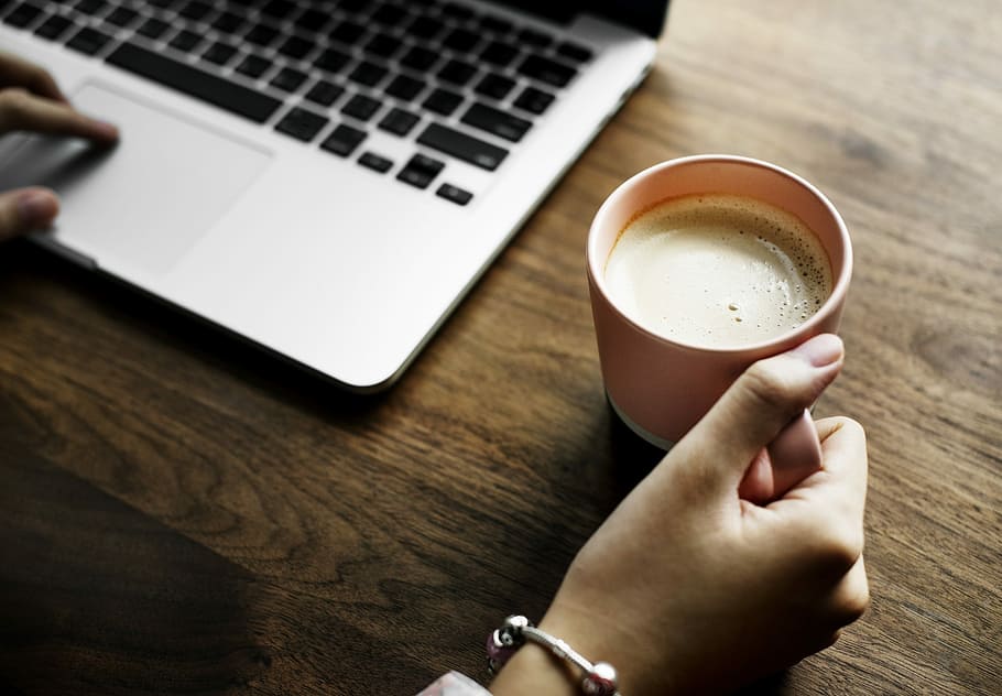 person holding mug while using MacBook, laptop, coffee, table