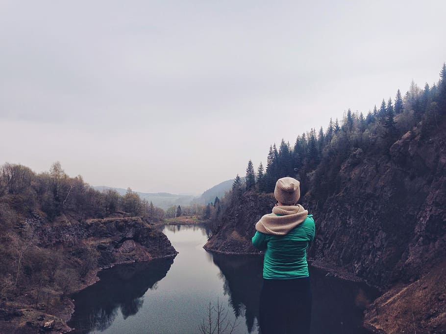 person wearing teal shirt with beige scarf and cap facing lake between mountains with forest HD wallpaper