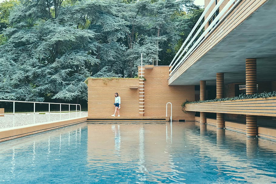 woman wearing white shirt standing beside brown bricked wall and blue water pool near trees during daytime, woman walking beside swimming pool near green leafed trees during daytime