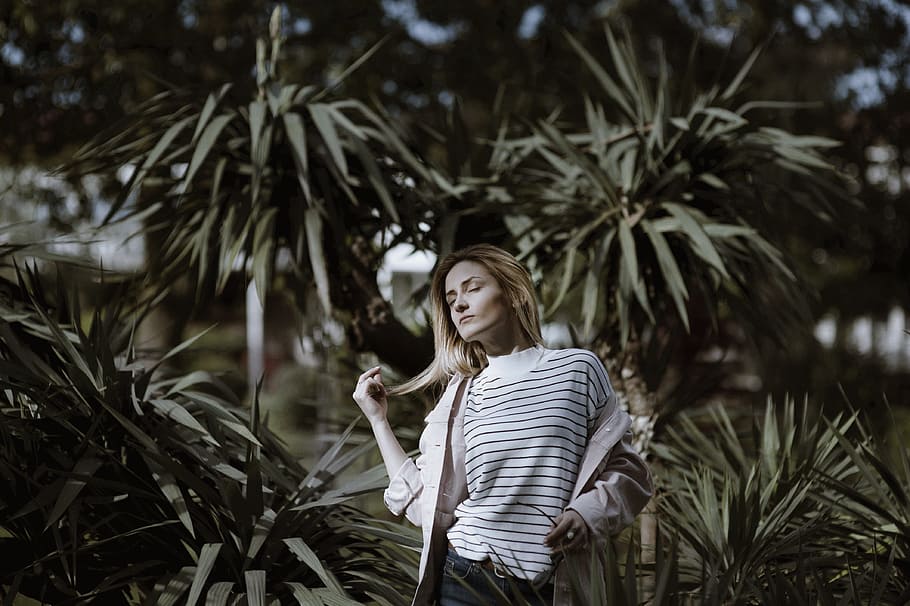 woman holding her hair closing eyes ner trees, woman standing beside green plant during daytime HD wallpaper