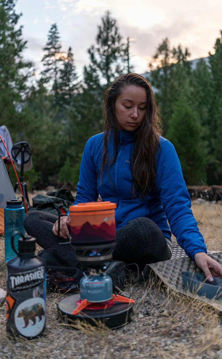 woman sitting on ground with camping stove and mat, woman squatting on ground beside camp gas stove