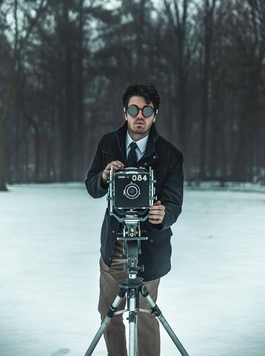 man standing in front of black camera with stand surrounded by trees, selective focus photography of man wearing black coat using black and grey camera with tripod near tall trees during daytime HD wallpaper
