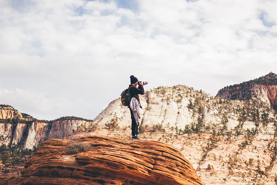 man wearing black jacket holding DSLR camera while taking photo standing on brown cliff during day time, person taking photo while standing on brown stone at daytime