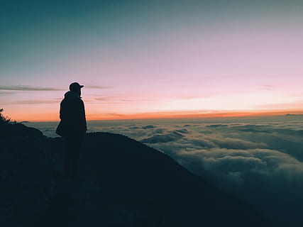 silhouette of man on hill facing horizon with orange sky, man standing on mountain top watching sea of clouds HD wallpaper