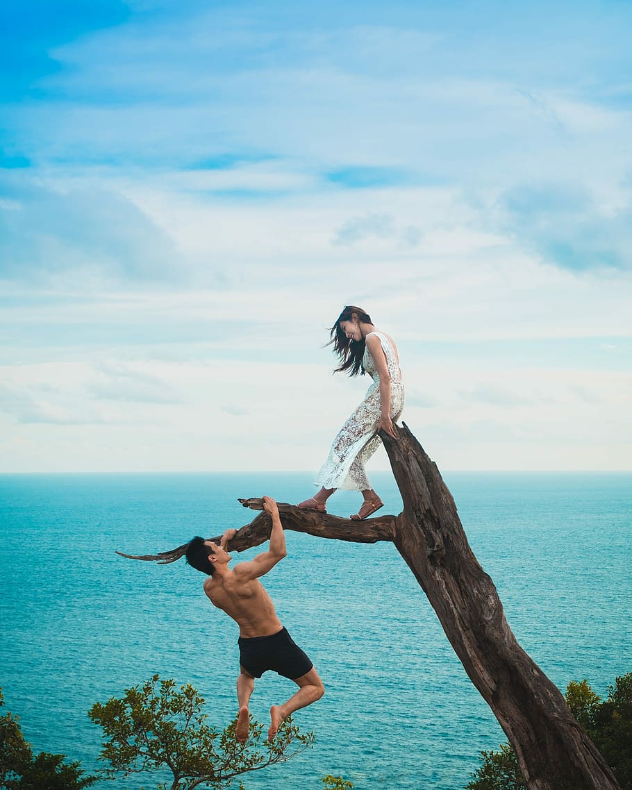 woman sitting on tree trunk with man holding on branch near sea under white clouds during daytime, woman on top of tree trunk and man climbing on it HD wallpaper