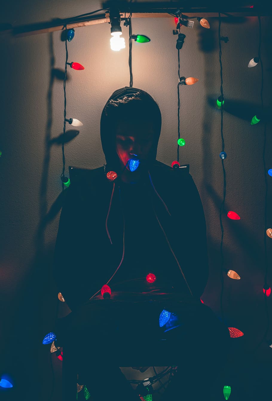 man sitting while biting multicolored string lights, person wearing black hoodie standing behind string lights, HD wallpaper