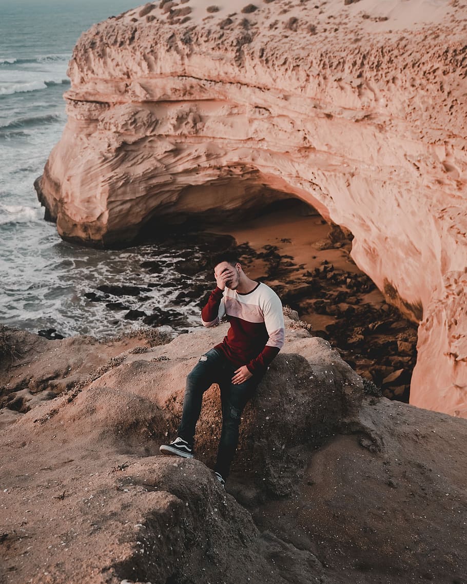 man covering face while sitting near the ocean water, man sitting on rock formation near sea during daytime HD wallpaper