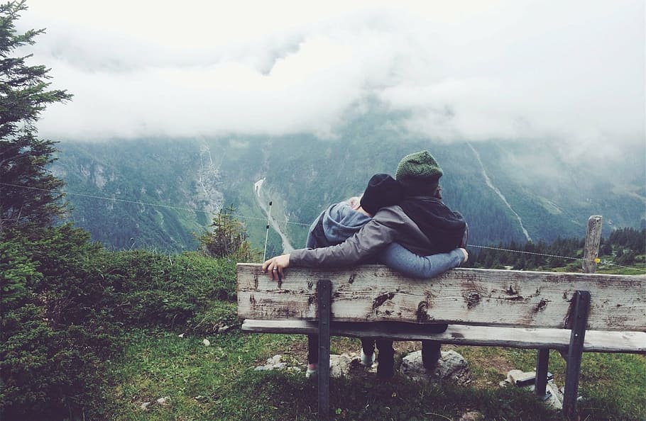 two person wearing jackets sitting on bench staring at mountain covered with trees and fog HD wallpaper