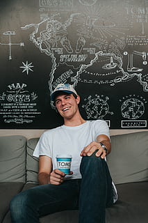 man sitting on couch while smiling, man sitting on gray couch wearing white crew-neck t-shirt holding TOMS cup over painted wall HD wallpaper
