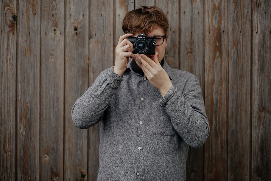 man holding black bridge camera taking photo and standing near brown wooden wall, man in  gray coat jacket a holding an DSLR camera HD wallpaper