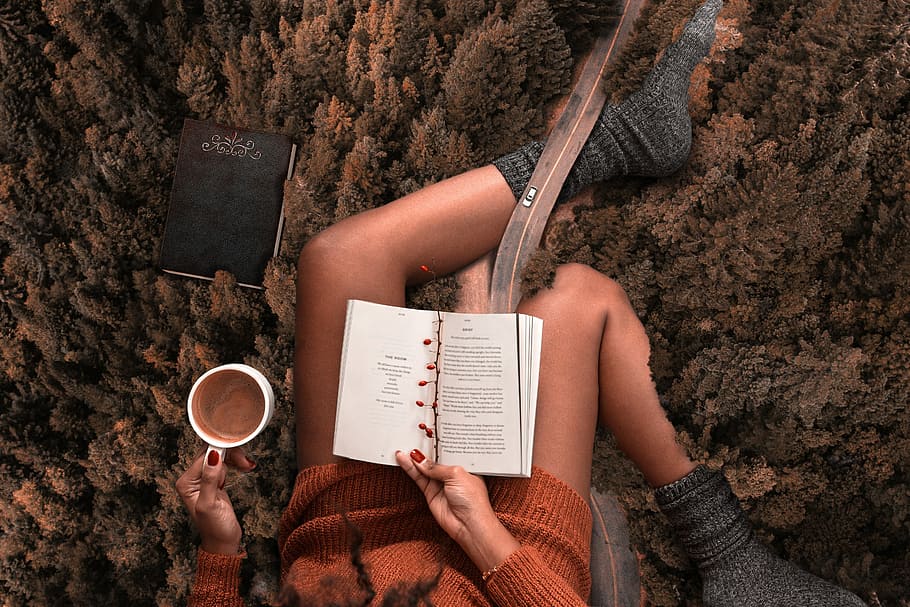 woman wearing orange sweater and grey socks reading a book with white mug