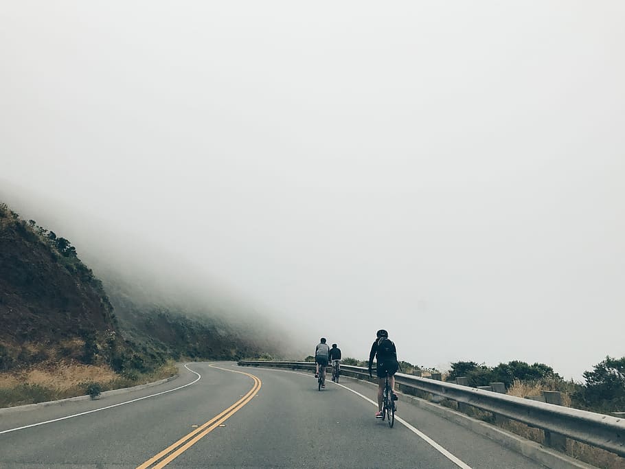 three men biking on asphalt road, three people biking on road near cliff with fog