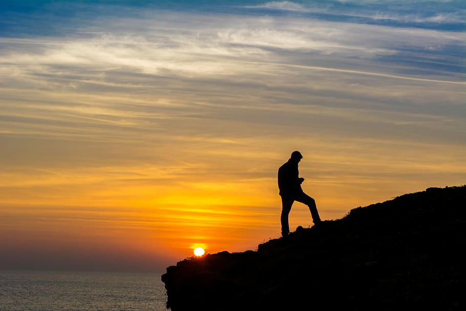 sihouette of person standing on rock formation during golden hour, silhouette of a man walking uphill during sunset