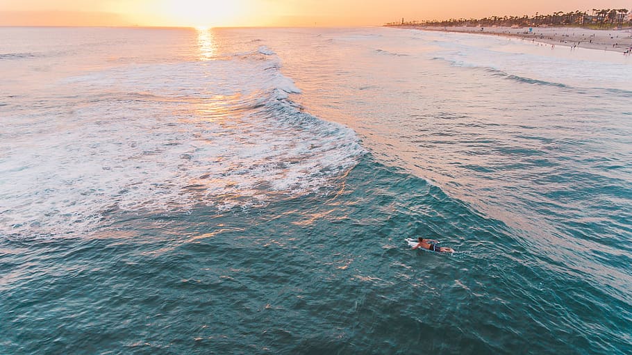 person riding on knee board on body of water, aerial photo of man wearing blue shorts swimming along with surfboard towards deep part of ocean near shore during daytime HD wallpaper