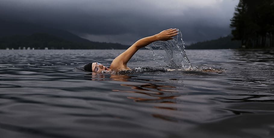 man swimming on body of water, person swimming on body of water surrounded with tall trees under cloudy sky