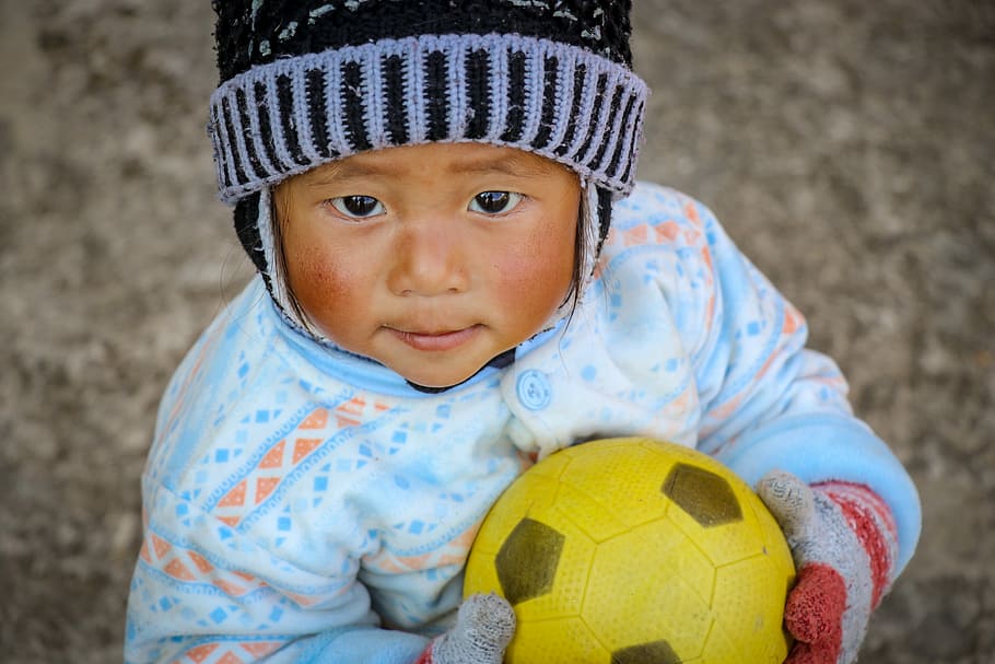 baby in blue and white shirt holding a ball, toddler wearing black and gray knit hat