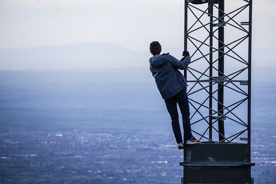 man climbing on tower near buildings at daytime, man in blue jacket stands on black tower overlooking city HD wallpaper