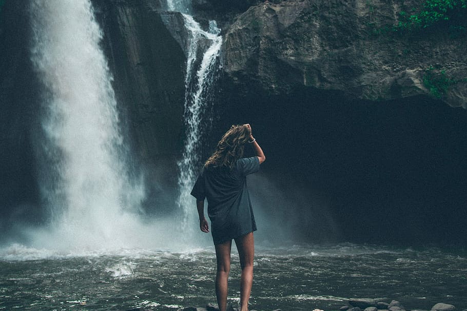 woman standing in front of waterfalls while holding her hair, woman standing in front of waterfalls during daytime