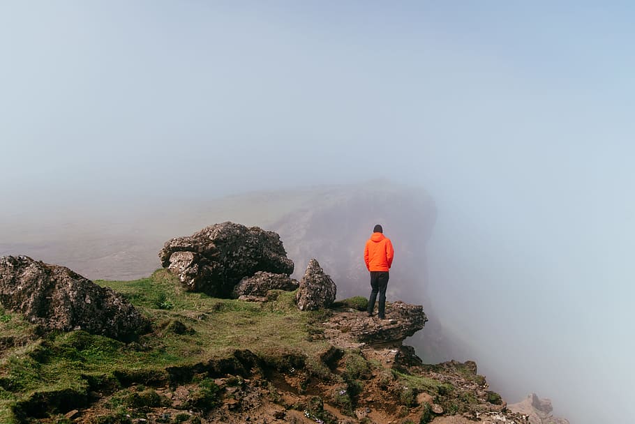 man wearing orange pullover hoodie, man standing on cliff during daytime HD wallpaper