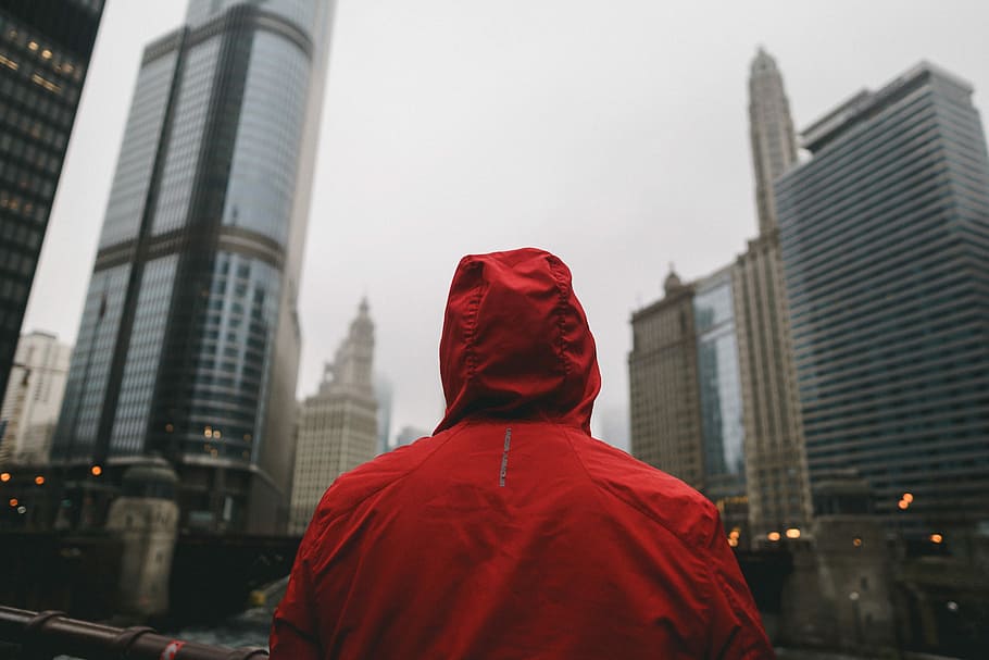 person wearing red hoodie standing in front of high rise buildings, person in red hooded jacket standing facing city buildings