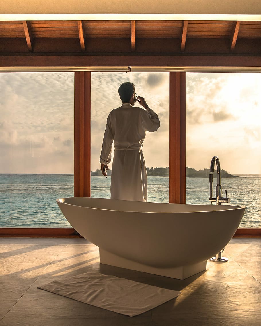 man standing beside bathtub, man wearing white bath robe standing next to free-standing bathtub inside bathroom near window during sunrise