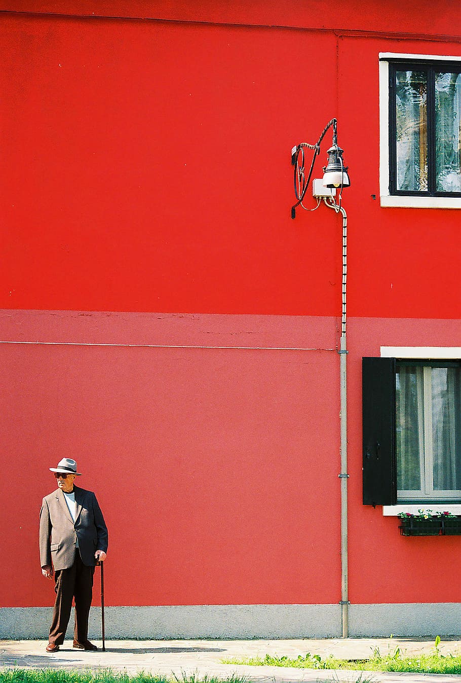 man wearing gray suit jacket and brown dress pants outfit holding cane standing in front of red and pink wall paint at daytime, man in suit standing near wall with lamp post HD wallpaper