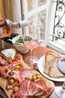 person pouring beverage on clear long-stem wine glass beside white ceramic plate with sliced bread and brown wooden board with bacons, person pouring wine glass beside bread and raw meat on table HD wallpaper