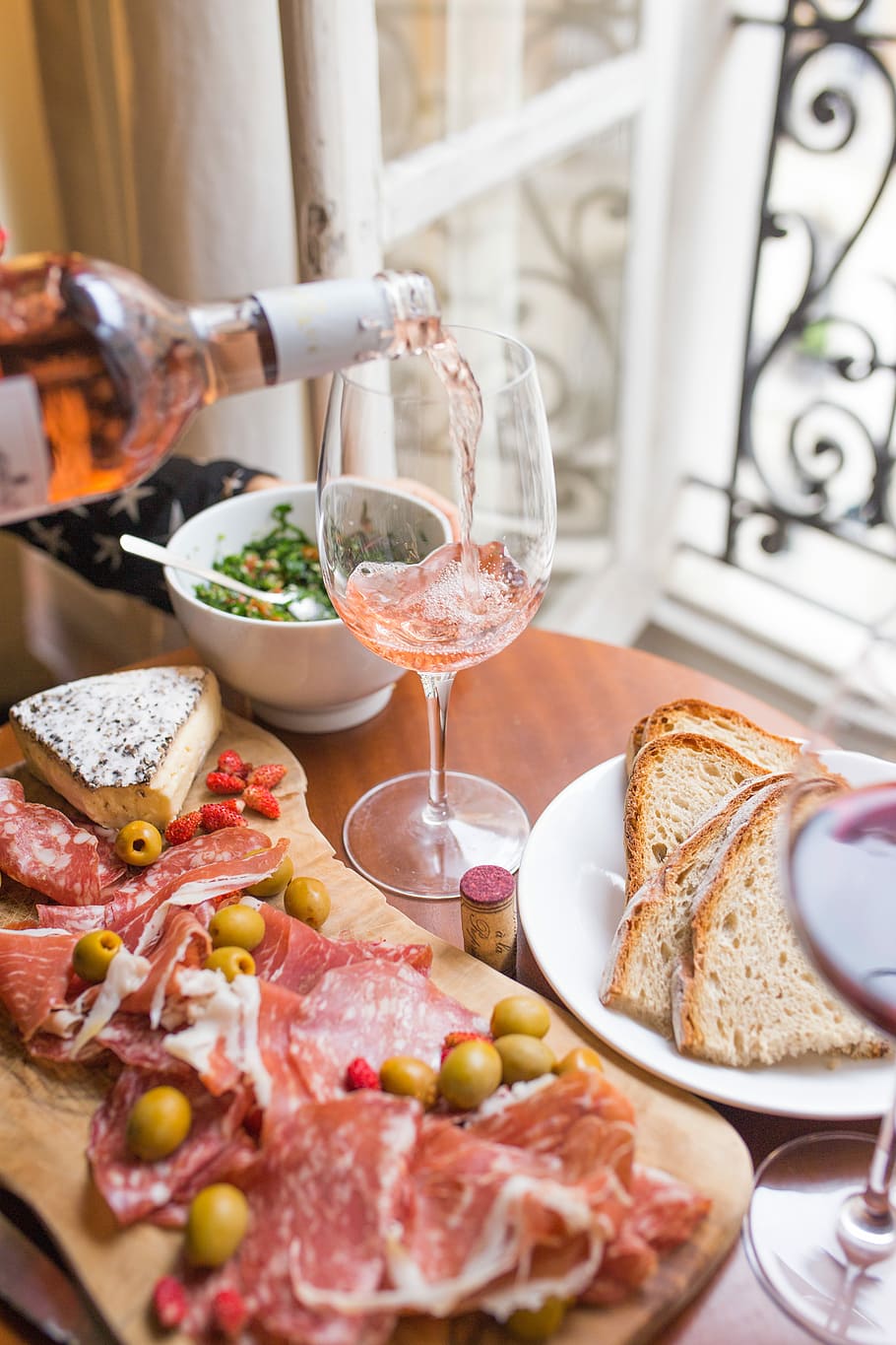person pouring beverage on clear long-stem wine glass beside white ceramic plate with sliced bread and brown wooden board with bacons, person pouring wine glass beside bread and raw meat on table HD wallpaper