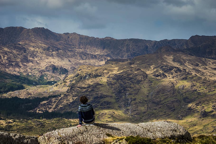 person sitting on concrete barrier overlooking mountain at daytime, person sitting on gray rock watching mountain HD wallpaper