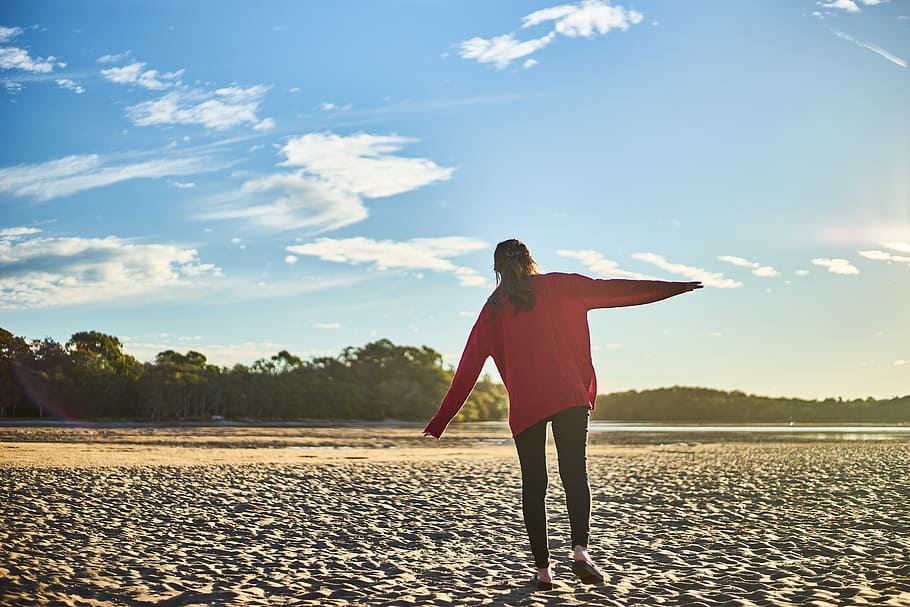 Woman in Red Sweater and Black Pants on White Sand Near Body of Water During Daytime HD wallpaper