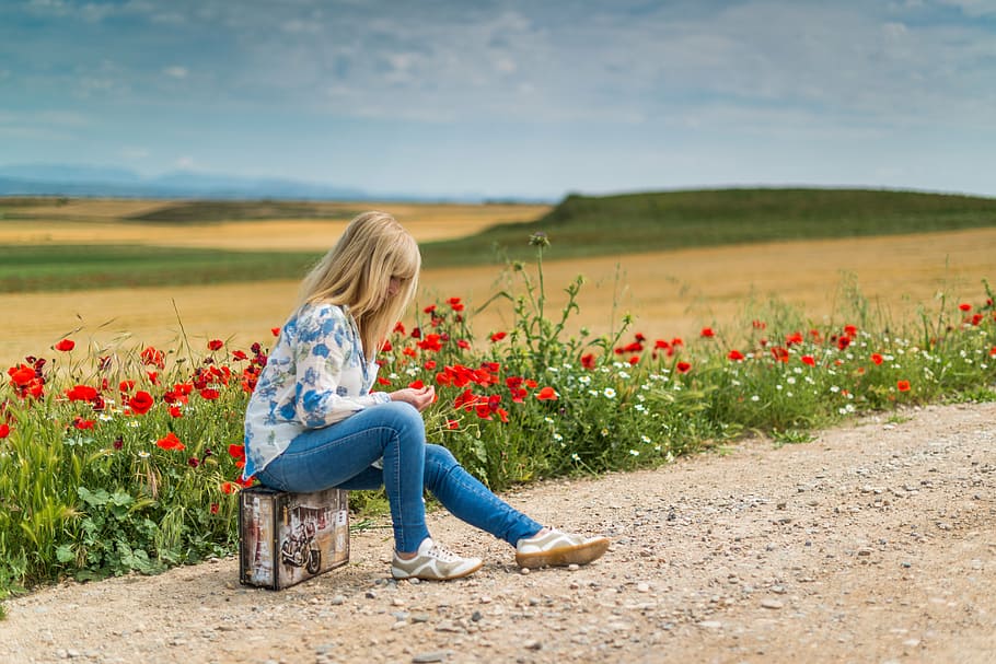 girl wearing blue and white floral long-sleeved shirt and blue fitted jeans sitting on briefcase beside red flowers during daytime, HD wallpaper