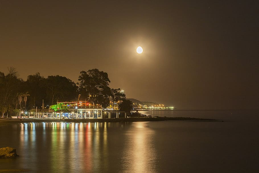 city lights near sea, white concrete building beside calm body of water at nightime HD wallpaper