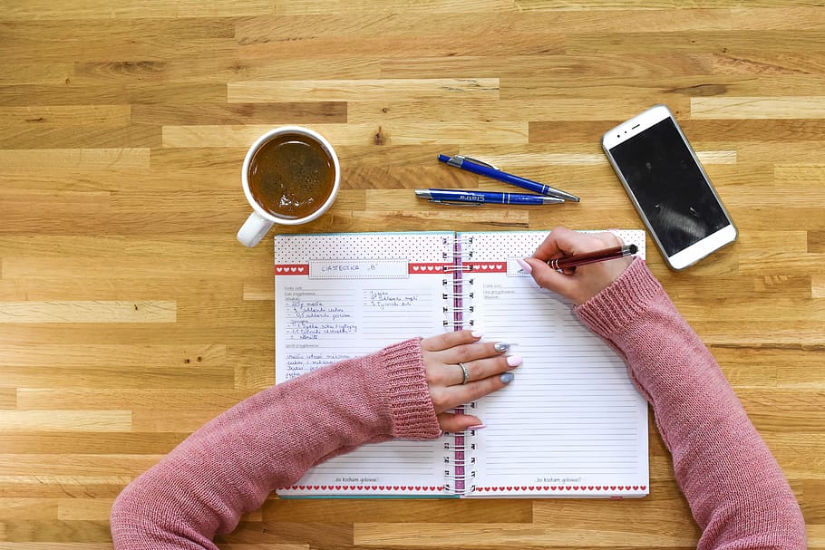 person writing on white notebook near white smartphone and cup on table HD wallpaper