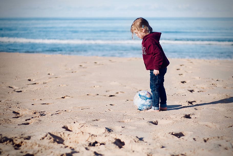 girl standing beside ball on seashore, child standing on beige sand near teal ball HD wallpaper