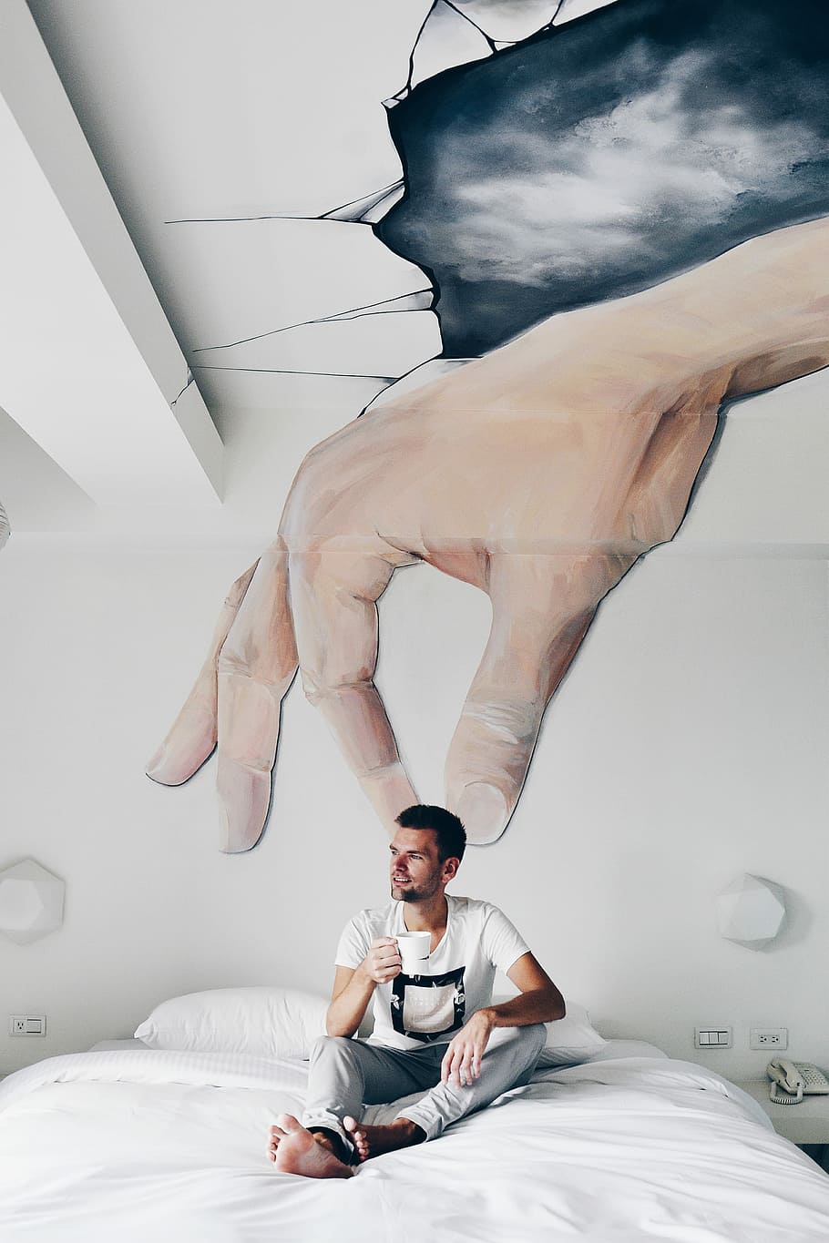 man sitting on bed while holding ceramic mug, man wearing white and black T-shirt during daytime HD wallpaper