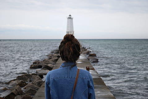 woman standing infront of lighthouse near beach, woman wearing blue denim top standing in front of white light post during daytime HD wallpaper