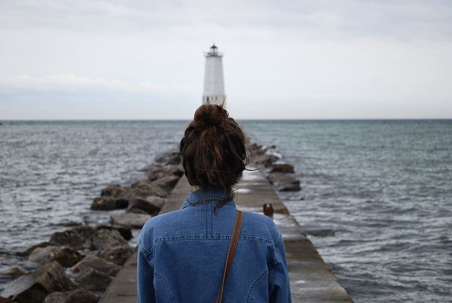 woman standing infront of lighthouse near beach, woman wearing blue denim top standing in front of white light post during daytime HD wallpaper