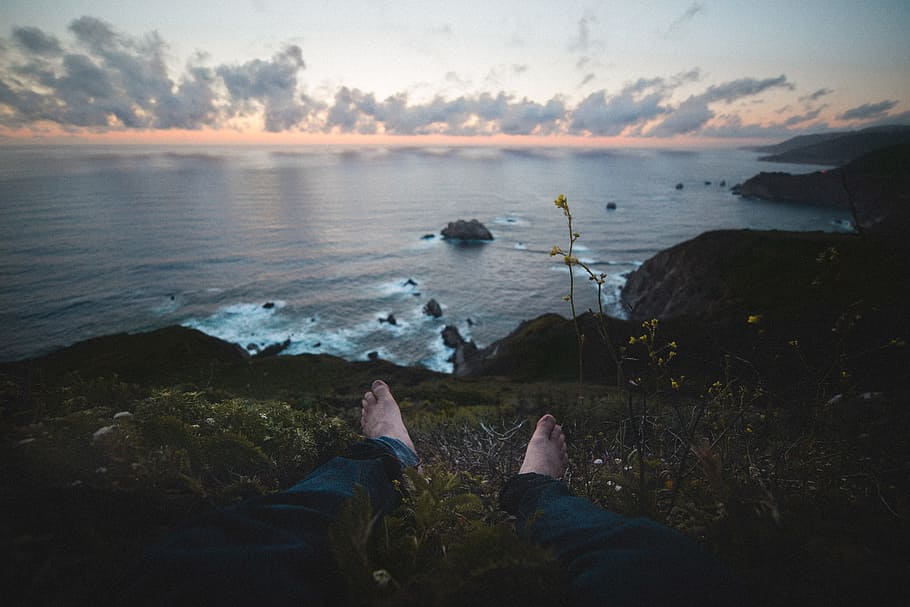 man sitting on green grass near body of water, person sits on grass at the peak front of sea at daytime HD wallpaper